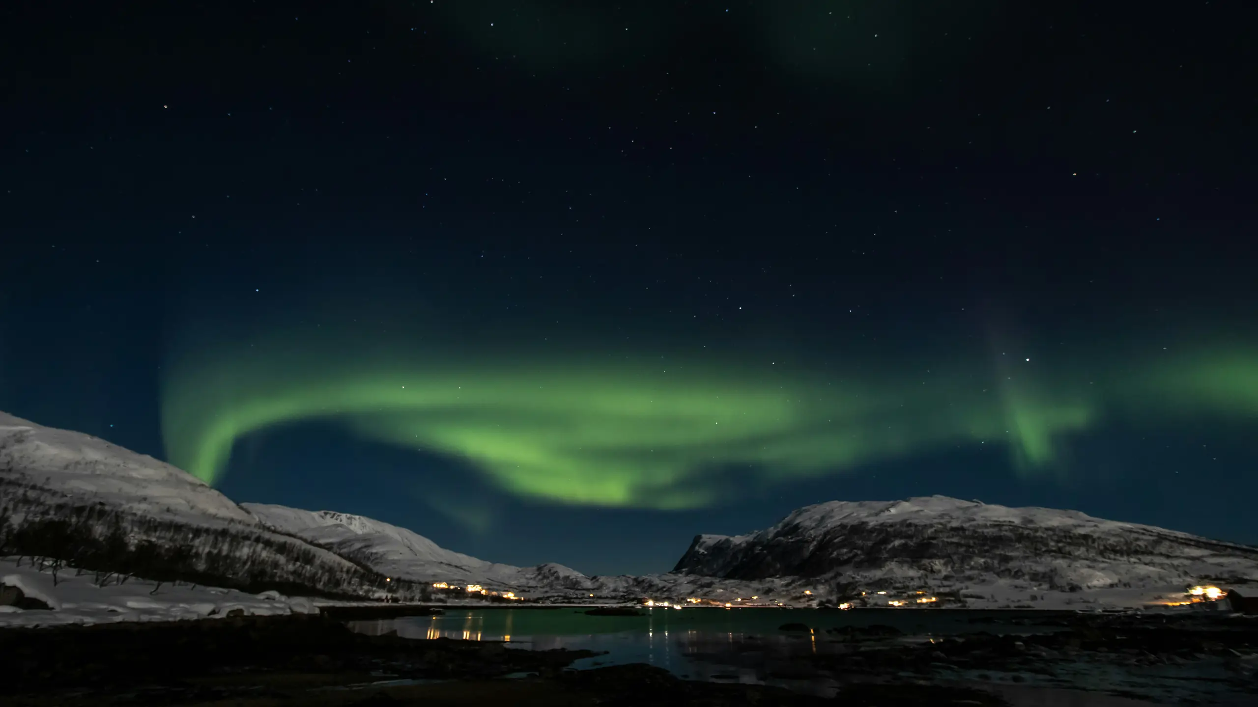 A green aurora arcing over the fjord at Ersfjordbotn outside Tromsø, with snow ridges on both sides and village lights at the waterline